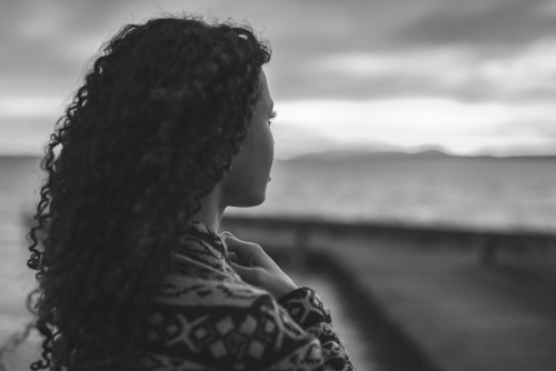 Young African American woman is standing on the promenade at the lake, looking thoughtfully towards the water and the setting sun. The girl in a vest is in a thoughtful mood