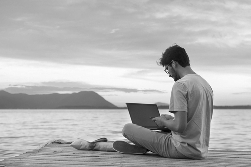 Digital nomad young man traveling and working remotely on laptop at a tropical island by the sea on the beach - Remote work outdoors and freelancer comcept.