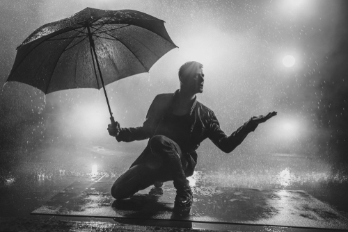 Young strange man with umbrella under the rain. Vibrant lights on background.