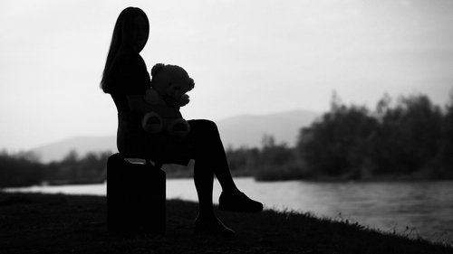 Silhouette of a girl sitting on a suitcase by the river. She is holding a teddy bear in her hands. Background illuminated by sunset light