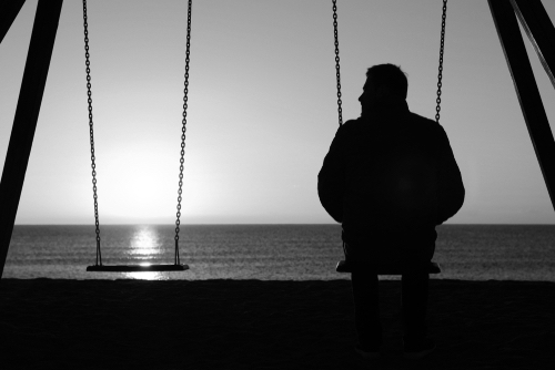 Man alone, back image, looking at the sunrise, siting on a swing, next to an empty swing. 