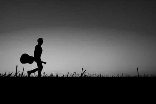 Silhouette of a male guitar player, holding his guitar, waling confidently on a path.