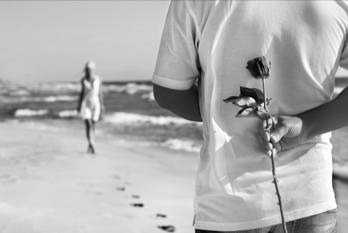 Man, at the shore on the beach, holding a rose on his back, for his griffins walking towards him. Waiting for her, uncertainty.