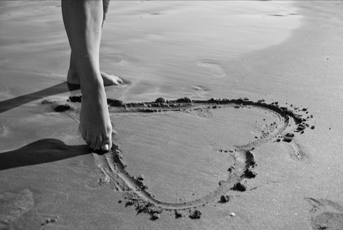 Woman's feet, drawing a heart on the sand with her toes: symbol of being ready for love, fragility, and vulnerability. 