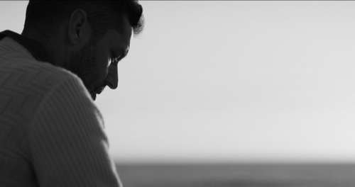 Man looking down, thoughtful, reflective, quiet presence - background peaceful beach and sky.