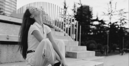 Female, sitting outdoors, next to a stair - with her had back, hair flowing down her back, closed eyes; image of letting go, release, relief, emotional expansion.