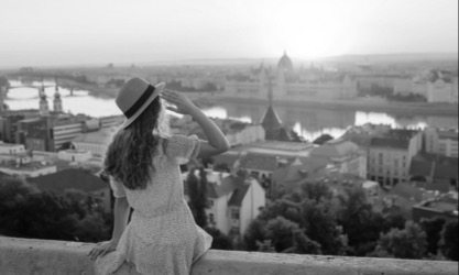 Female, sitting up high, on a fence edge, her hand above her eye washing the city and the sunrise. Image of healing, transformation, new life, emotional freedom, and possibilities.