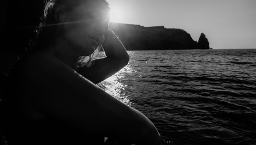 Women, seating at the beach - the coat mountains and a sun ray in the background, she is gazing toward the shoreline. Image of stillness, introspection, resilience, healing and hope. 